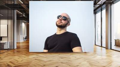 Confident young Caucasian man wearing sunglasses and a black T-shirt, standing outdoors against the sky. Handsome bald male model in shades thinking of success in the city Wall mural