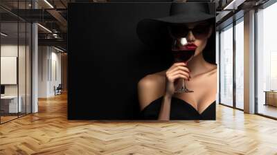 A stylish woman in a black hat and sunglasses holds a glass of red wine against a dark background. Wall mural