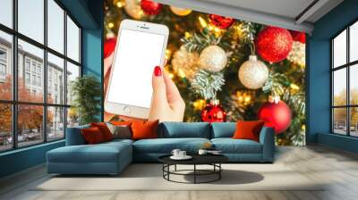 A woman's hand displays a blank smartphone in front of a beautifully decorated Christmas tree adorned with shimmering lights Wall mural