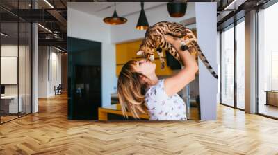 cheerful woman raises bengal cat up standing in the kitchen Wall mural