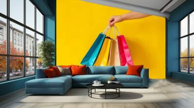 Hand holding colorful shopping bags against an upbeat yellow background suggesting retail and consumerism Wall mural