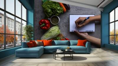 Cropped shot of hand writing recipes on book with food ingredients and fresh vegetables in the kitchen, top view Wall mural