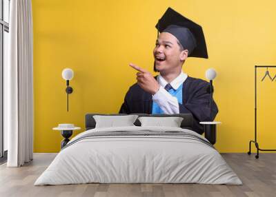 Excited Asian male in graduation clothes looking and pointing aside to the copy space for text or ads. Wall mural