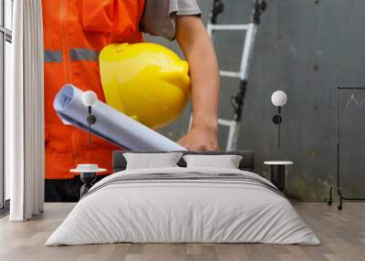 Front view of male construction worker holding blueprint and safety helmet outdoor of construction site with ladder in the background. Copy space and selective focus. Wall mural