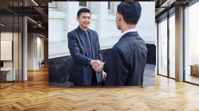 Two smiling businessmen shaking hands together while standing outside office building. Asian businessman shake hands with his colleague, over shoulder view Wall mural