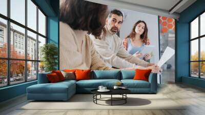 A business team engages in a serious conversation with expressions of focus and concern in a brightly lit, contemporary office space, providing a glimpse into corporate dynamics. Wall mural