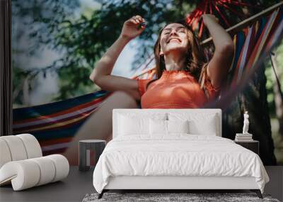 A joyful woman in a red top relaxes in a colorful hammock under a tree, enjoying a sunny summer day. Wall mural