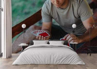 A man skillfully holds a fan of playing cards at an outdoor wooden table, showcasing his concentration and enjoyment in the leisure activity. Wall mural