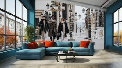 A multicultural group of young entrepreneurs confidently strides through a cityscape, engaged in a remote working session or business meeting. Wall mural