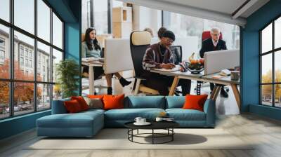 A multicultural office scene with focused employees working at desks. Men and women of various ages demonstrate teamwork and professionalism in a contemporary workspace. Wall mural