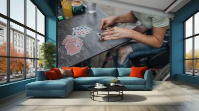 A person in casual attire concentrates on playing cards at a table with drinks. The scene is relaxed, highlighting leisure and social interaction. Wall mural