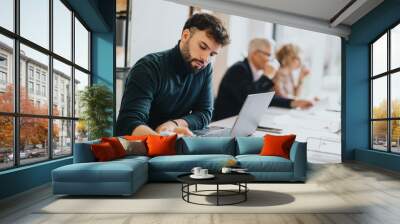 A young man diligently working on his laptop in a busy office setting, representing focus and professionalism in the workplace. Wall mural