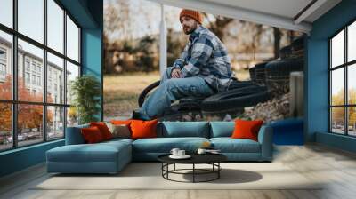 A young man in casual attire sits leisurely on stacked used tires, taking in the tranquility of a sunny park setting. Wall mural