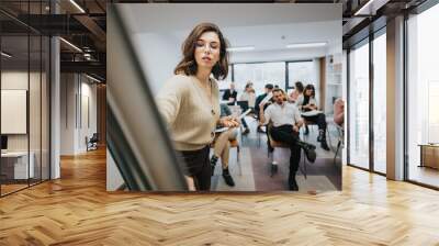 A young woman leads a presentation at a business workshop, confidently engaging with a group of professionals seated in a stylish office setting. Wall mural