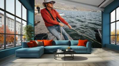 An adult male in casual attire pulls a rope to secure a boat by a beautiful lake with mountains and boats in the distance. Wall mural