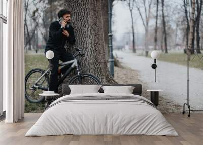 An adult man in casual business attire stops his bike ride in a serene park to have a conversation on his smart phone, balancing work and health. Wall mural