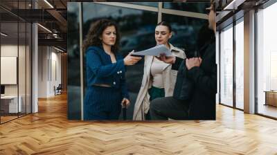 Business colleagues engage in discussion while reviewing documents outside a modern office building. Professional attire and serious expressions indicate a work-related conversation. Wall mural