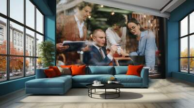 Business meeting of four colleagues analyzing reports, brainstorming strategies and discussing revenue growth at a modern cafe using laptops and documents. Wall mural