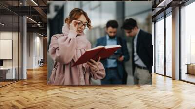 Focused female professional reading a notebook with male colleagues discussing in the background, portraying teamwork and collaboration outdoors. Wall mural