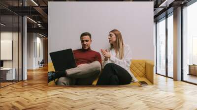 Good looking male person showing something on the lap top to his female colleague. They are taking a break from work and sitting on yellow sofa at modern co working space Wall mural