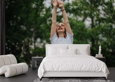 Happy woman in a white shirt joyfully stretches her arms upwards amidst a vibrant green park. She exudes positivity and freedom in this natural outdoor setting. Wall mural