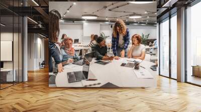 Long curly haired male employee helping his colleagues with financial charts while working with them in modern office room Wall mural