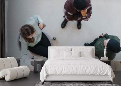 Overhead shot of a relaxed business meeting with three professionals engaging over a laptop. One female and two males in casual office attire converse in a modern workspace setting. Wall mural