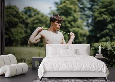 Shirtless young man using a resistance band for outdoor exercise in a lush green park on a bright, sunny day. Wall mural