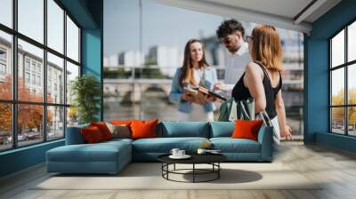 Three young professionals engage in a business discussion near a river in a modern city. They are reviewing documents and enjoying a sunny day outside. Wall mural