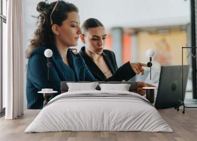 Two businesswomen discuss and collaborate on a project, focusing on a laptop for work in a modern office setting. Wall mural