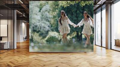 Two carefree young females dressed in lovely white dresses running hand in hand enjoying the sunshine and freedom in a verdant park during springtime. Wall mural