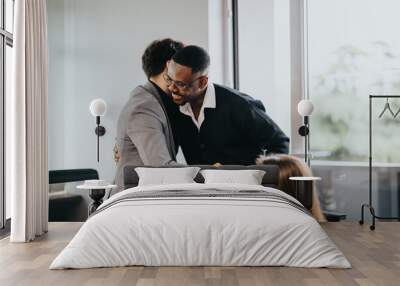 Two male business colleagues embrace in a well-lit office, showing support and camaraderie. A female coworker watches, contributing to the welcoming and inclusive work culture depicted. Wall mural