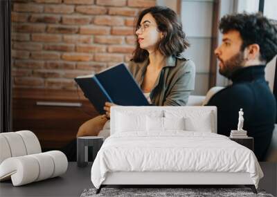 Two professionals in a casual office setting, woman reading a book with interest, man in contemplation, warm brick backdrop. Wall mural