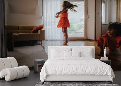 Two young girls enjoying a playful moment indoors, dancing and moving in a bright, sunny living room with wooden floors and a sofa. Wall mural