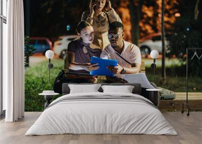 Young adults collaborating on a project at night, using a digital tablet. They are focused on work while a colleague talks on the phone, surrounded by evening city lights. Wall mural