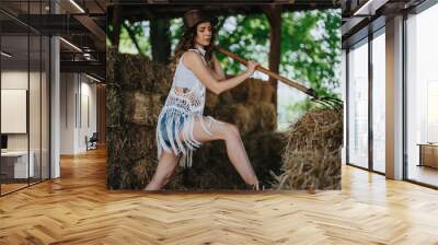 Young female farmer in a barn, working with hay. She is wearing casual summer attire and a hat, conveying hard work and rural life. Wall mural