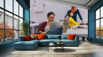 Young woman sitting at her table in the office with her legs on the desk while working on her laptop.. Wall mural