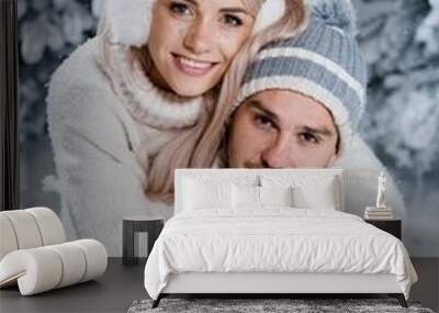 New year love story. Couple kisses and hugs. People weared wearing fur headphones, hats, white sweaters. Happy young couple hugs and kiss near christmas trees in winter day. Wall mural