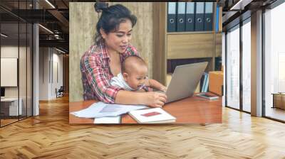 Pretty young single mom working at home on a laptop computer while holding her baby girl sitting on her lap enjoying playing computer. Wall mural