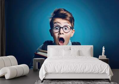 Surprised little boy in glasses with backpack reading book on blue background. Back to school concept. Wall mural