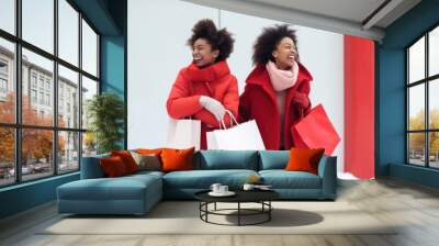 Two cheerful female friends holding shopping bags on snowy winter day. Diversity women making shopping during Christmas sales season. Wall mural