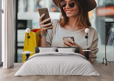 A beautiful young woman in a fashionable outfit, using her smartphone and smiling as she holds shopping bags.  Wall mural