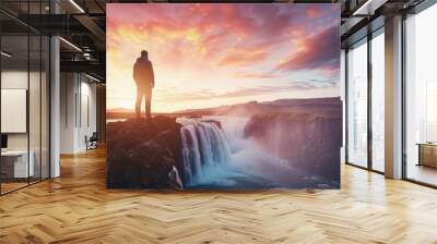 GoÃ°afoss waterfall flows peacefully with a colorful sunset sky overhead. A male tourist in casual attire stands on a cliff, taking in the beautiful summer scenery. Wall mural