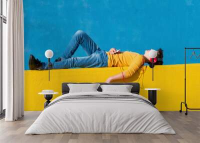 Front view of a young boy wearing casual clothes lying on a yellow fence against a blue wall while using a mobile phone to listening music by headphones Wall mural