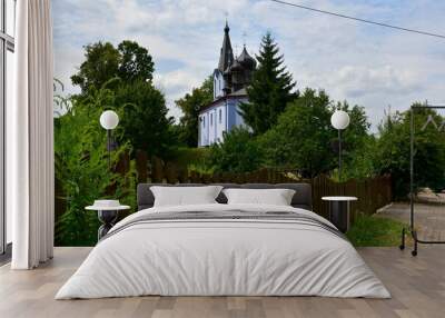 A close up on an old blue Catholic or Orthodox church located behind a wooden fence and surrounded with lush flora, including trees, shrubs, flowers, and herbs seen on a cloudy summer day in Poland Wall mural