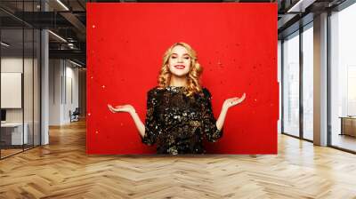 Holiday and party concept: beautiful young woman wearing evening dress standing under confetti rain over red background Wall mural