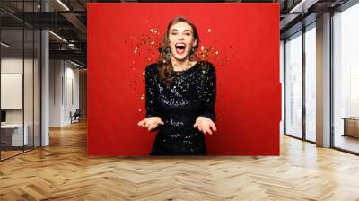 Holiday concept: beautiful brunette female wearing evening dress standing under confetti rain over red background Wall mural