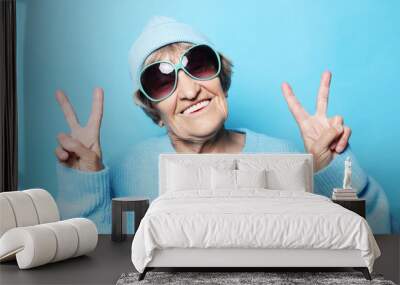 Lifestyle, emotion and people concept: Funny old lady wearing blue sweater, hat and sunglasses showing victory sign. Wall mural