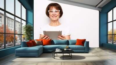 Lifestyle, tehnology and old people concept: An elderly woman in a white T-shirt and glasses is holding an iPad and smiling. Wall mural