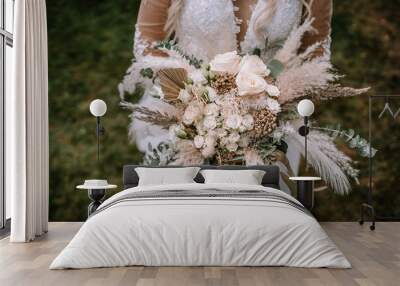 A bride is holding a large, ornate bouquet featuring roses, pampas grass, and various greenery, focused on the flowers with the bride's lace gown in the background. Wall mural